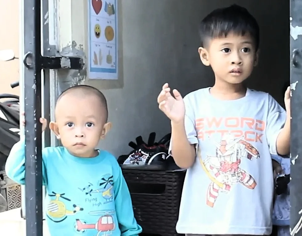 Dua anak laki-laki DaycareQu berdiri di pintu pagar; yang lebih besar mengangkat tangan dan melambai.