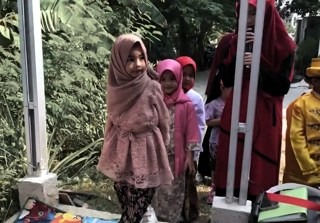 Seorang anak perempuan berkerudung dan berbaju brokat merah muda berdiri di depan teman-temannya yang berbaris di belakang.