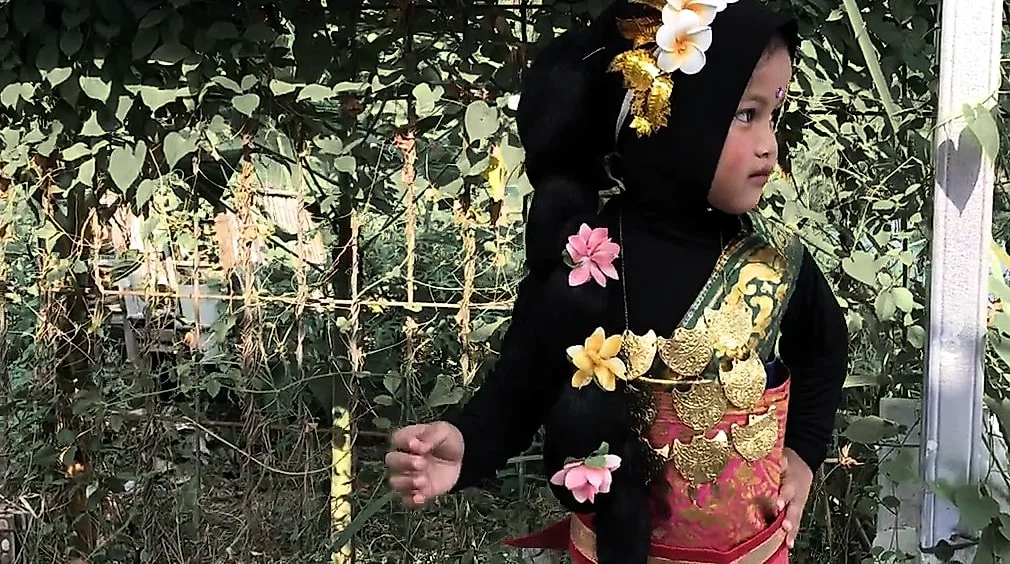 Seorang anak perempuan berkerudung hitam dengan busana adat, hiasan bunga kamboja di kepala dan hiasan emas di dada, berdiri di depan pagar tanaman.