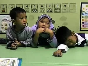 Tiga anak RA di meja kelas: seorang anak laki-laki menyimak, anak perempuan di sebelahnya menopang dagu, dan seorang lagi merebahkan kepala di meja.