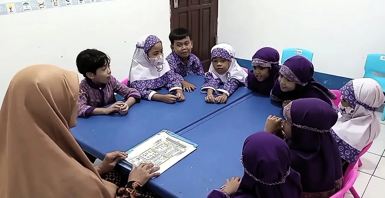 Seorang guru berkerudung cokelat duduk membelakangi kamera di meja biru, selembar kertas di depannya; di seberangnya, anak-anak RA berseragam ungu menyimak.