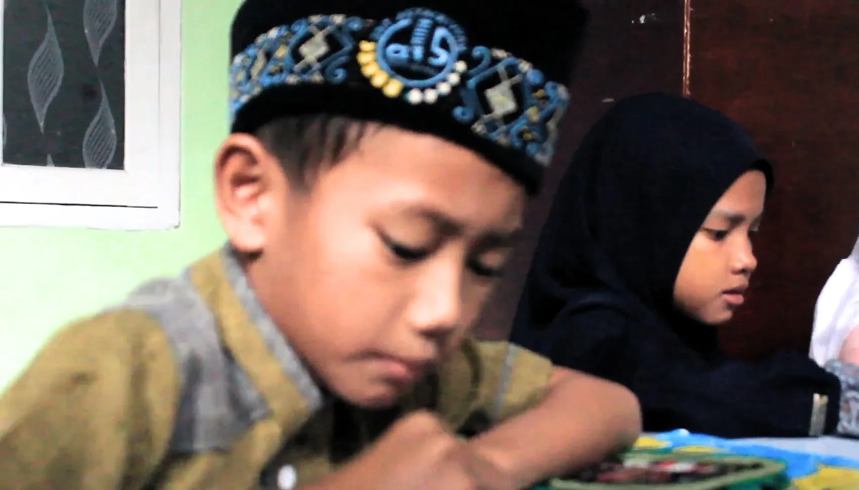 Seorang anak laki-laki berpeci dan dua anak perempuan berkerudung menunduk, menulis di meja kelas.