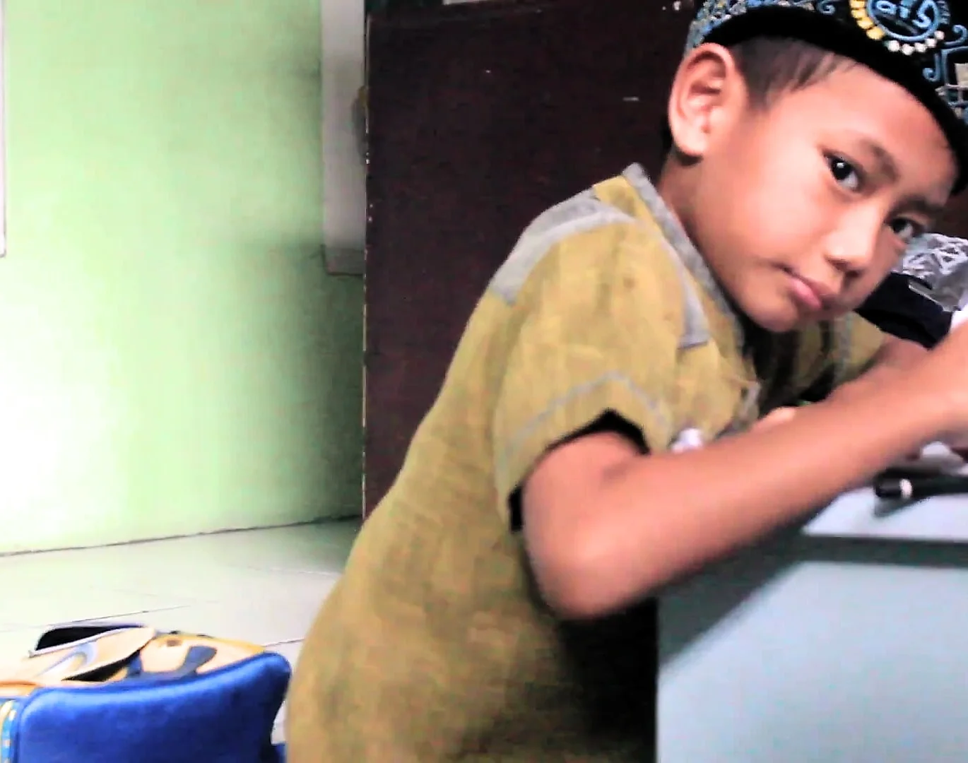 Seorang anak laki-laki berpeci menulis di meja kelas dan menoleh ke arah kamera; di belakangnya dinding hijau muda.