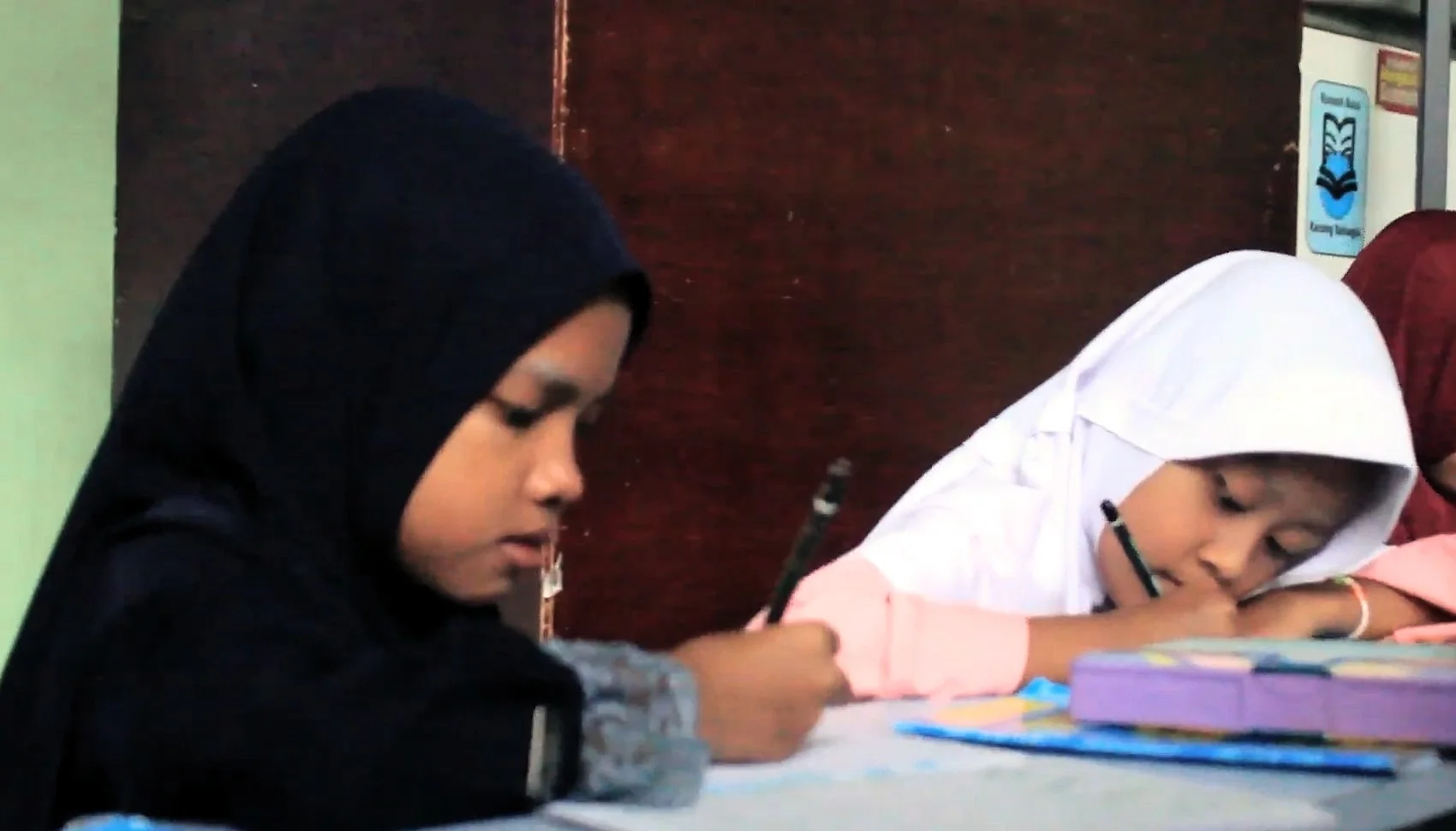 Anak-anak perempuan berkerudung gelap, putih, dan merah marun menulis di meja; sebuah kotak pensil ungu di depan mereka.