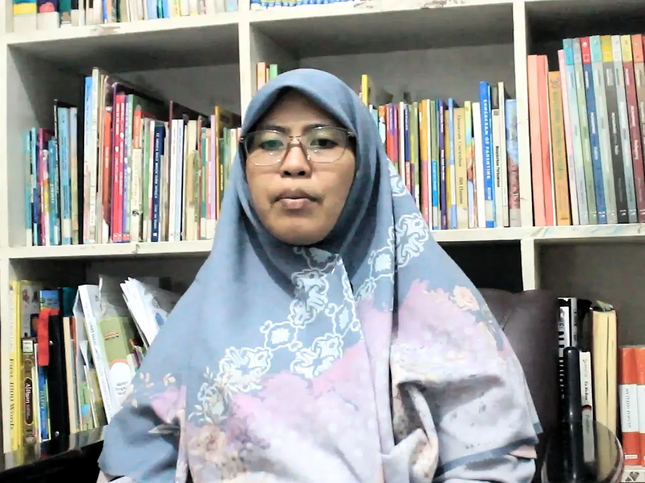 Seorang perempuan berkerudung biru keabu-abuan dan berkacamata duduk di depan rak buku.