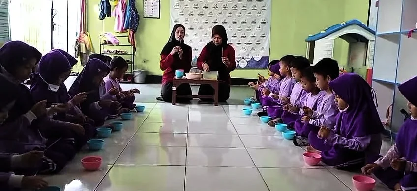 Anak-anak DaycareQu duduk berjajar di lantai di sepanjang dinding, dengan mangkuk di depan masing-masing; dua orang dewasa duduk di balik meja kecil sambil memegang sate buah.