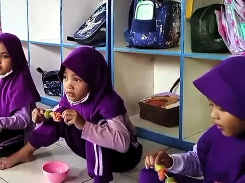 Seorang anak perempuan DaycareQu berkerudung ungu memegang sate buah, duduk di lantai di depan rak tas.