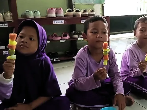 Anak-anak DaycareQu duduk di lantai, masing-masing memegang sate buah warna-warni.