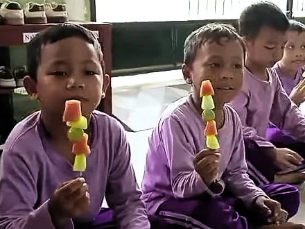 Dua anak laki-laki DaycareQu berkaus ungu tersenyum ke kamera sambil memegang sate buah.