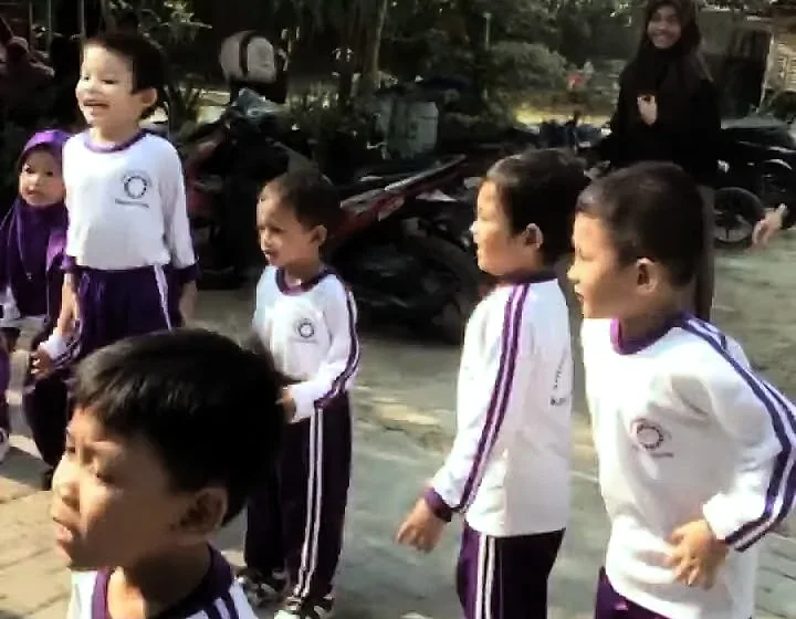 Anak-anak RA berseragam olahraga putih-ungu berbaris di jalan berpaving; seorang anak di depan tertawa.