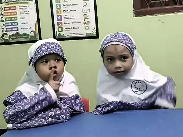 Dua anak perempuan RA berkerudung duduk berdampingan di meja biru, menyimak ke arah depan kelas.