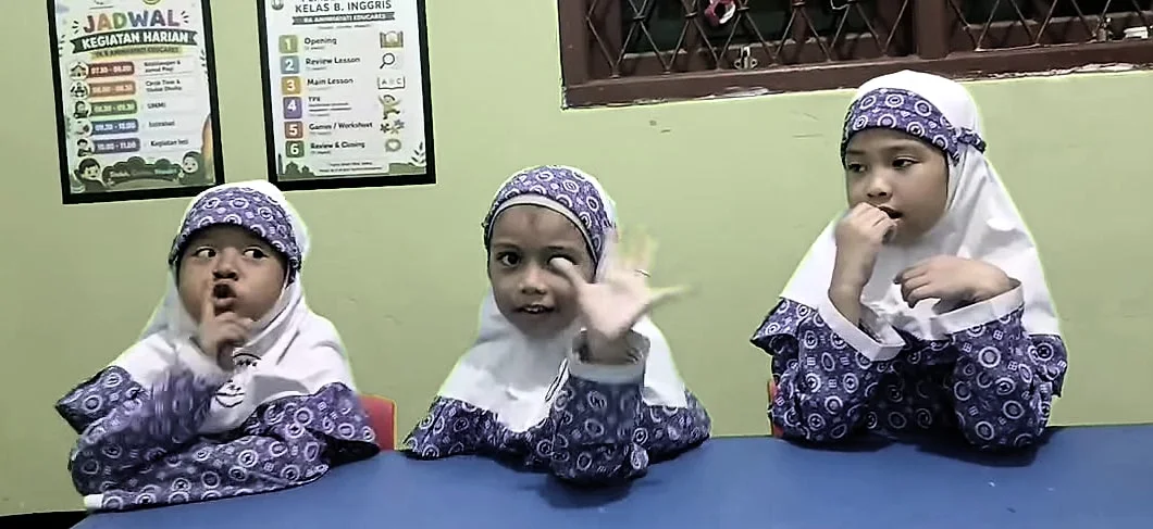 Tiga anak perempuan RA berkerudung duduk berjajar di meja biru; yang di tengah melambaikan tangan.